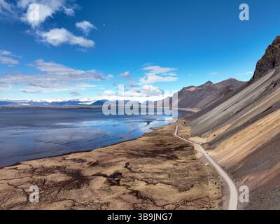 Strada tortuosa sulla costa meridionale dell'Islanda che traccia il bordo di una buia e ancora baia, dove le aspre montagne incontrano una costa arida e ventosa sotto un vibran Foto Stock