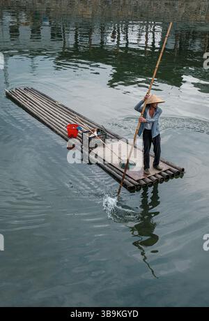 Barcaiolo che indossa un cappello di paglia pagaiando una zattera di bambù sull'acqua verde del fiume a Jingdezhen, Jiangxi, Cina Foto Stock