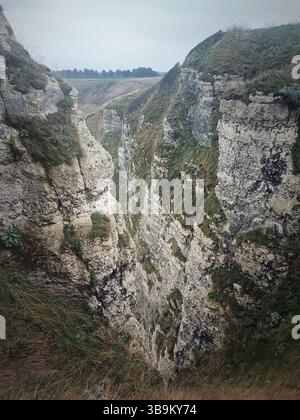 Una gola profonda e stretta con ripide scogliere rocciose ricoperte da macchie di erba e vegetazione. Il paesaggio è aspro e suggestivo, con una vista distante di Foto Stock