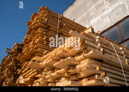 Tavole di legno disposte in modo ordinato sono impilate accanto a una vecchia parete di mattoni, asciugando con luce naturale. Una cinghia verticale in plastica tiene saldamente la struttura in p Foto Stock
