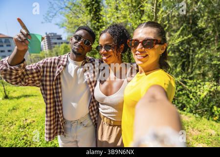 Tre felici amici multietnici che fanno un selfie in un parco, uno di loro sta puntando il dito verso il cielo Foto Stock