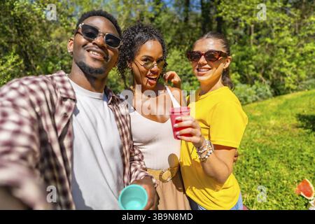 Tre amici allegri si godono un picnic, si scattano un selfie e tengono in mano tazze colorate in una giornata di sole nel parco Foto Stock