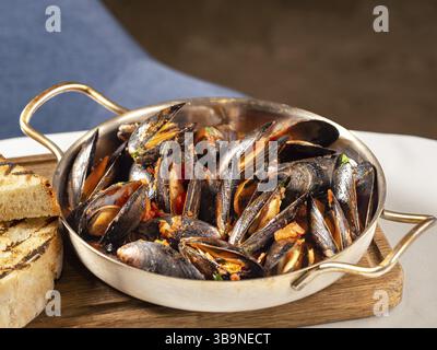 Deliziose cozze nere in salsa di pomodoro rosso. Cozze nere mediterranee in teglia metallica su tagliere in legno con pane alla griglia sul tavolo del ristorante b Foto Stock