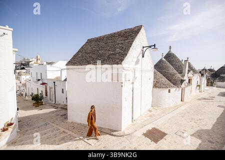 Donna che cammina lungo un'affascinante strada acciottolata ad Alberobello, circondata da iconiche case di trulli imbiancate con tetti conici sotto un cielo azzurro Foto Stock