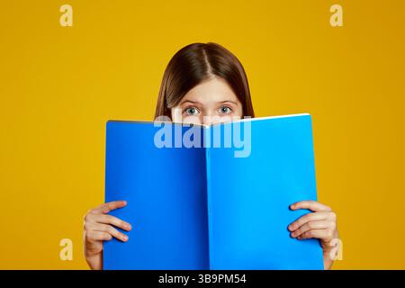 Studente curioso che tiene in mano una cartella blu, che sbircia con occhi giocosi su uno sfondo giallo brillante Foto Stock