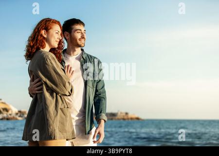 Una giovane coppia si trova in riva al mare sorridente e avvolgente mentre guarda in lontananza, godendosi un momento di sole tranquillo vicino all'acqua. LIF Foto Stock