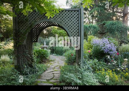 Sentiero in pietra ammiraglia attraverso un mandrino in legno delimitato da arbusti di Kerria japonica «Picta» e mauve Campanula lactiflora «varietà di Prichard» in estate. Foto Stock