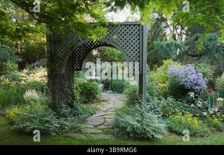 Sentiero in pietra ammiraglia attraverso un mandrino in legno delimitato da arbusti di Kerria japonica «Picta» e mauve Campanula lactiflora «varietà di Prichard» in estate. Foto Stock