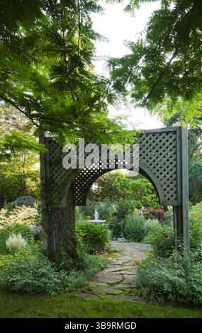 Sentiero in pietra ammiraglia attraverso un mandrino in legno a reticolo delimitato da arbusti di Kerria japonica «Picta» nel giardino del cortile in estate. Foto Stock