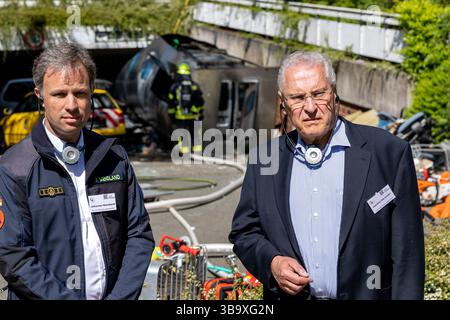 10 maggio 2025, Baviera, Erlangen: Il ministro degli interni della Baviera Joachim Herrmann (l, CSU) ascolta le spiegazioni dell'esercizio di controllo delle catastrofi "Löwenzahn" in un sito di emergenza tramite cuffie insieme a Johannes Wendland, capo della filiale di Erlangen dell'Agenzia federale tedesca per il soccorso tecnico (THW). Nello scenario di esercitazione, un aereo cargo si è schiantato sopra il centro della città. Parti di un alto edificio sono state gravemente danneggiate. I piani superiori di un edificio di uffici adiacente sono in fiamme. E in un impianto chimico fittizio vicino, la coda dell'aereo schiantata ha danneggiato diversi contenitori con Foto Stock
