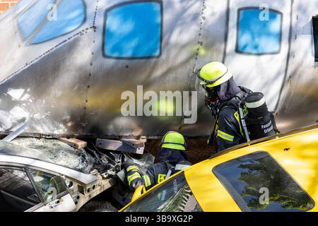 10 maggio 2025, Baviera, Erlangen: I vigili del fuoco stanno lavorando su un cockpit (modello) di aerei che si è schiantato in un vialetto di parcheggio sotterraneo come parte dell'esercitazione di controllo delle catastrofi "Löwenzahn". Nello scenario di esercitazione, un aereo cargo si è schiantato sopra il centro della città. Parti di un alto edificio sono state gravemente danneggiate. I piani superiori di un edificio di uffici adiacente sono in fiamme. E in un impianto chimico fittizio nelle vicinanze, la coda dell'aereo schiantata ha danneggiato diversi contenitori con merci pericolose. La cabina di pilotaggio dell'aereo si è schiantata su un'auto all'ingresso di un parcheggio sotterraneo Foto Stock
