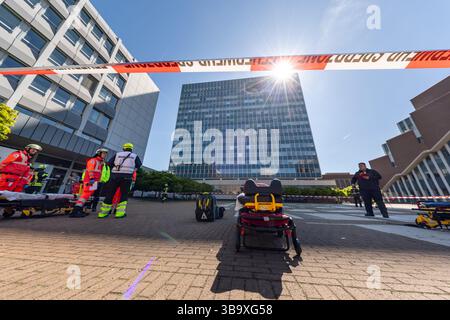 10 maggio 2025, Baviera, Erlangen: I servizi di emergenza conferiscono di fronte a un edificio in cui un aereo si è schiantato nell'ambito dell'esercitazione di controllo delle calamità "Löwenzahn" (nella foto su un telone nelle file superiori delle finestre). Nello scenario di esercitazione, un aereo cargo si è schiantato sopra il centro della città. Parti di un alto edificio sono state gravemente danneggiate. I piani superiori di un edificio di uffici adiacente sono in fiamme. E in un impianto chimico fittizio nelle vicinanze, la coda dell'aereo schiantata ha danneggiato diversi contenitori con merci pericolose. La cabina di pilotaggio dell'aereo si è schiantata contro un'auto all'ingresso di un unde Foto Stock