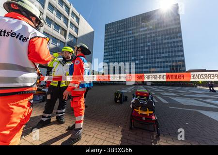 10 maggio 2025, Baviera, Erlangen: I servizi di emergenza conferiscono di fronte a un edificio in cui un aereo si è schiantato nell'ambito dell'esercitazione di controllo delle calamità "Löwenzahn" (nella foto su un telone nelle file superiori delle finestre). Nello scenario di esercitazione, un aereo cargo si è schiantato sopra il centro della città. Parti di un alto edificio sono state gravemente danneggiate. I piani superiori di un edificio di uffici adiacente sono in fiamme. E in un impianto chimico fittizio nelle vicinanze, la coda dell'aereo schiantata ha danneggiato diversi contenitori con merci pericolose. La cabina di pilotaggio dell'aereo si è schiantata contro un'auto all'ingresso di un unde Foto Stock