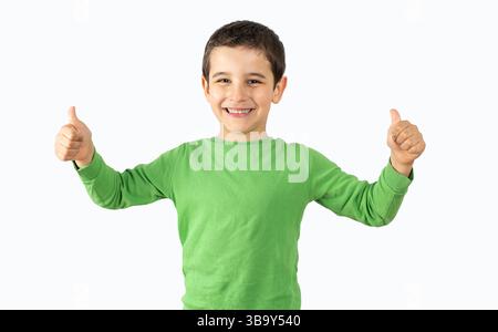 Foto in studio di un bambino che indossa una t-shirt casual verde su sfondo bianco con un cartello di successo che fa un gesto positivo con la mano, con i pollici sorridenti e felici. Foto Stock