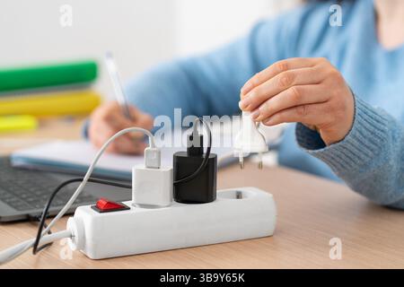 Connettore maschio per la mano di una studentessa su una presa multipla che utilizza un computer portatile su una scrivania a casa Foto Stock