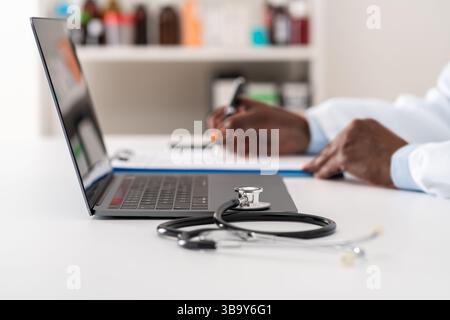 Primo piano di uno stetoscopio appoggiato su un computer portatile con un medico irriconoscibile che lavora in background Foto Stock