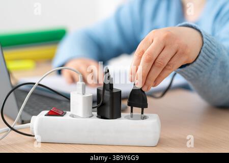 Primo piano di una studentessa che collega la spina a una presa multipla utilizzando un computer portatile su una scrivania di casa Foto Stock