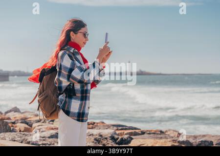 Giovane donna con zaino e sciarpa rossa che scatta foto del paesaggio marino con il faro utilizzando lo smartphone Foto Stock