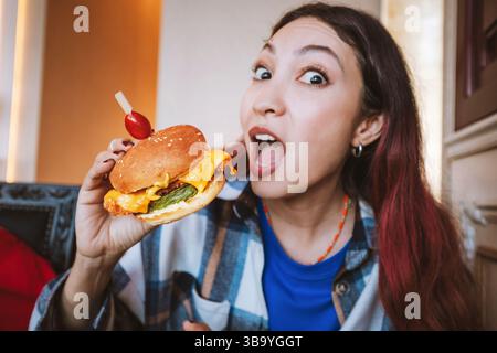 Giovane donna eccitata che tiene in mano un gustoso hamburger e apre la bocca, pronta a mangiare, in un ristorante Foto Stock