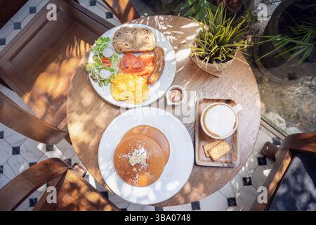 Una deliziosa colazione e' presentata su un tavolo di legno caratterizzato da pile di soffici pancake conditi con burro, una porzione di uova strapazzate, salsiccia e un Foto Stock