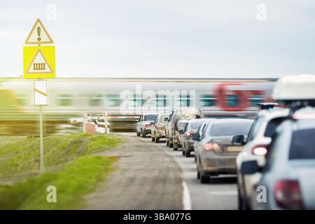 Una coda di auto aspetta che un treno passeggeri viaggi in un passaggio ferroviario fuori città. La barriera automatica impedisce l'accesso non autorizzato Foto Stock