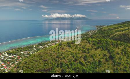 La lussureggiante foresta tropicale copre il terreno collinare di Tahiti, le case colorate del villaggio lungo la costa, l'isola di Moorea all'orizzonte sotto il cielo nuvoloso. Natura remota, esotici viaggi estivi. Ripresa aerea Foto Stock
