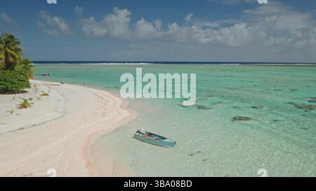 L'acqua turchese della laguna incontra la spiaggia di sabbia rosa in una giornata di sole con poche nuvole e piccole onde sullo sfondo, con una barca ancorata in primo piano, ad Aitutaki, Isole Cook Foto Stock