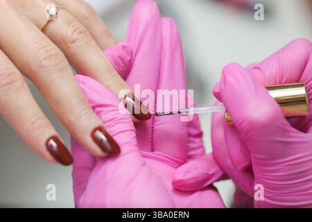 Procedura di applicazione dello smalto per unghie. Manicure con guanti rosa. Salone di bellezza per la cura delle mani. Pennello di precisione a colori. Trattamento cosmetico professionale. Foto Stock