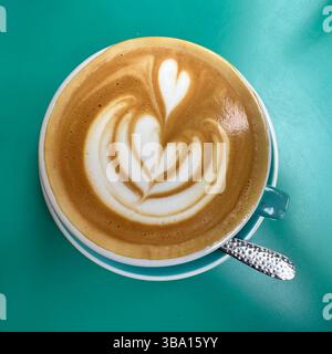 Caffè bianco piatto con cuore latte art servito in una tazza di tè su un tavolo turchese. Brunch luminoso in stile minimalista. Foto Stock