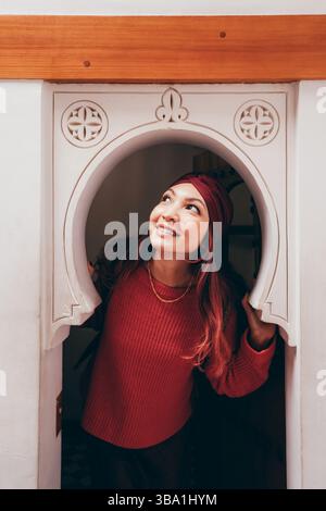Giovane donna con un velo che guarda in alto e sorride mentre esplora la tradizionale architettura marocchina Foto Stock