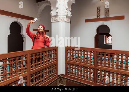 Giovane donna che scatta selfie con lo smartphone nel cortile del riad, circondato dalla tradizionale architettura marocchina Foto Stock