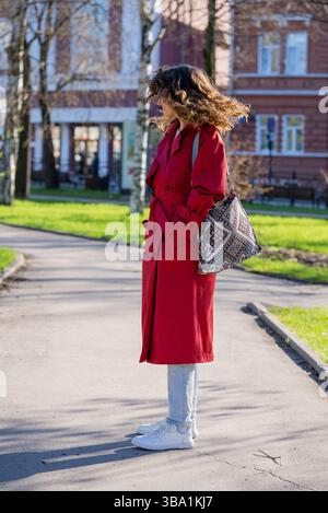 Donna elegante con capelli ricci, con un cappotto rosso e scarpe da ginnastica bianche, cammina in un parco cittadino in una giornata di primavera assolata, godendosi l'aria fresca e l'ambiente urbano Foto Stock