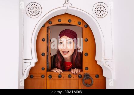 Giovane turista che indossa un turbante che sbircia da una porta tradizionale marocchina a Marrakech Foto Stock