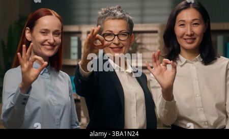 Tre donne d'affari multietniche co-lavoratrici caucasiche asiatiche team donne raccomandano femmine che mostrano gesto ok ok ok sorridenti colleghi approvazione ufficio Foto Stock