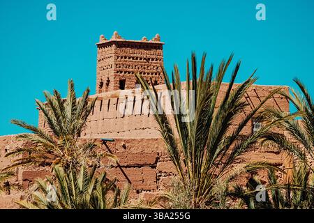 Palme che ricoprono parzialmente una kasbah marocchina fatta di mattoni di argilla sotto un cielo blu profondo Foto Stock