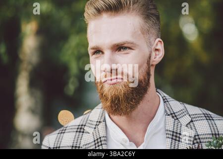 Bell'uomo barbuto in abiti vintage. Ritratti lo sposo elegante in una giacca a quadri il giorno del suo matrimonio nel parco. Ritratto hipster rustico. Uomo barbuto. Foto Stock