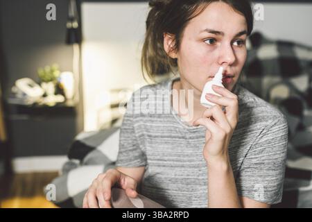 Il tema è stagionale freddo, naso che cola virus influenzale infezione. Giovane donna caucasica a casa camera da letto letto utilizza spruzzare gocce di medicina nel suo naso per sno Foto Stock