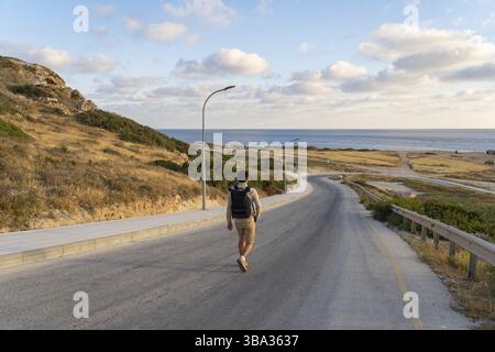 Uomo turistico con zaino passeggiate in montagna lungo strada asfaltata verso il mediterraneo a cipro nella zona di Agios Georgios Pegeias. Vista posteriore di b Foto Stock