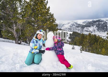 Famiglia, madre e figlia, pupazzo di neve con foresta e catena montuosa sullo sfondo. Vacanze invernali sugli sci ad Andorra, Pirenei Foto Stock