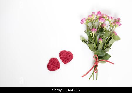 Rose rosa fresche con nastro rosso e due cuore rosso all'uncinetto. Disposizione piatta su sfondo bianco, vista dall'alto Foto Stock