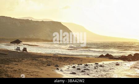 Due sdraio su una spiaggia selvaggia vuota illuminata da un tramonto dorato. Piccole onde sulla roccia, colline sullo sfondo nebbioso, Lapta, Cipro, Europa Foto Stock