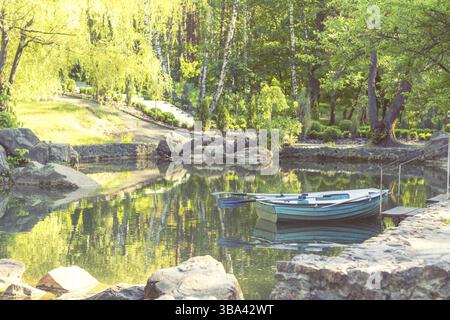 Soleggiata giornata primaverile nel parco cittadino con uno stagno circondato da rocce. Gli alberi riflettono nell'acqua. Barca di legno blu con due pagaie che riposa in pace Foto Stock