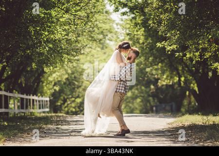Lo sposo felice solleva la sposa nel parco estivo. l'uomo ha cresciuto e abbracciato la donna alle sue braccia. Coppia sposata di recente nel parco. Solo sposato. Cammina nel parco An Foto Stock