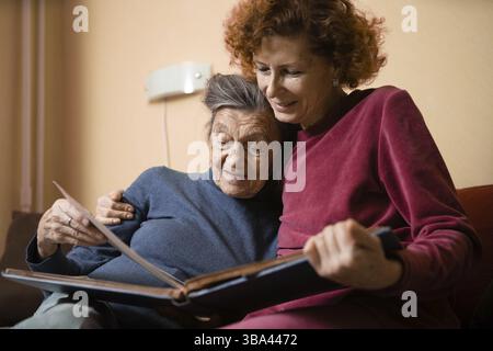 La figlia matura visita la madre anziana e ha divertimento guardando album fotografico di famiglia, seduto in abbraccio nel divano del soggiorno. Festa delle madri. Donna anziana e. Foto Stock