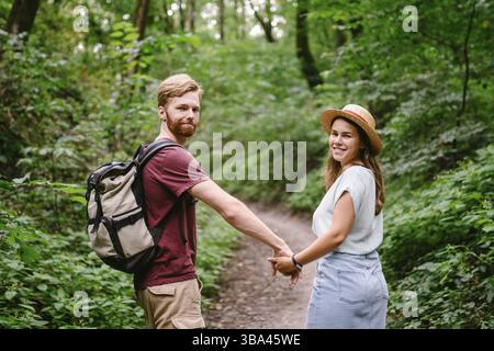 Coppia che si tiene per mano camminando nella foresta, vista posteriore. Avventura, viaggi, turismo, escursioni, gente. Vista posteriore di due persone che trasportano lo zaino mentre camminano Foto Stock