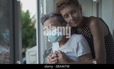 Figlia matura che visita la mamma senior in casa di cura. La figlia anziana abbraccia la sua madre di 90 anni in maschera medica mentre si trova alla finestra in blocco Foto Stock