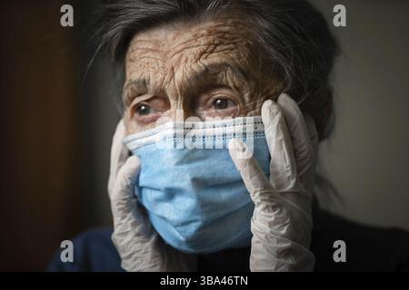 Ritratto donna anziana che tiene le mani della testa, triste emozione preoccupata in maschera medica protettiva, guanti contro virus e infezione a casa. Vecchi capelli grigi fe Foto Stock