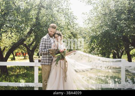 Coppia caucasica innamorata sposa e sposo in piedi in abbraccio vicino a una recinzione rurale bianca in legno in un parco di mele. il tema e' il ritratto di nozze e l'essere Foto Stock