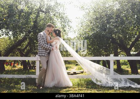 Coppia caucasica innamorata sposa e sposo in piedi in abbraccio vicino a una recinzione rurale bianca in legno in un parco di mele. il tema e' il ritratto di nozze e l'essere Foto Stock