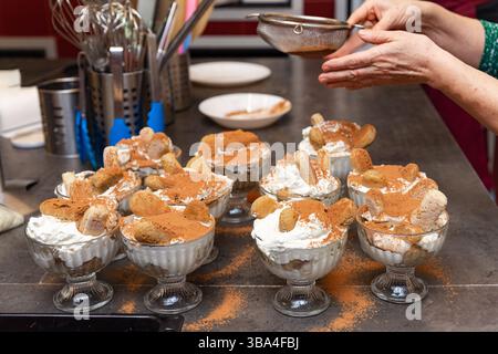 Più tazze in vetro di tiramisù fatti in casa sono ricoperte di cacao in polvere utilizzando un setaccio in un ambiente accogliente in cucina. Foto Stock